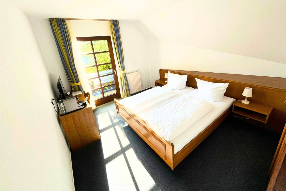 A hotel room with a large bed, a wooden headboard, a bedside table with a lamp, and a window with curtains.
