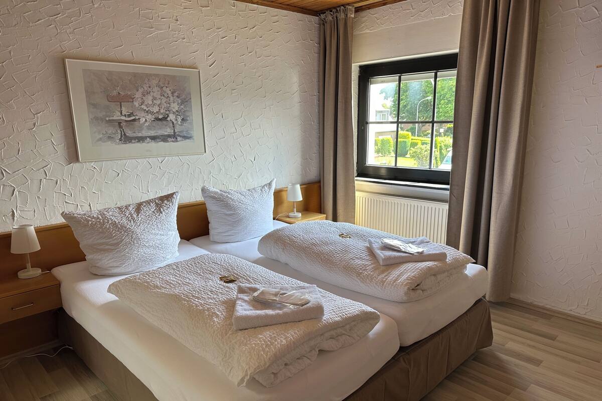 A neatly made double bed with white linens and towels, a framed picture on the wall, and a window with curtains.
