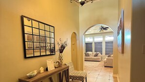 A hallway with a wooden console table, a mirror, and a chandelier. There is a chair and a couch in the background.