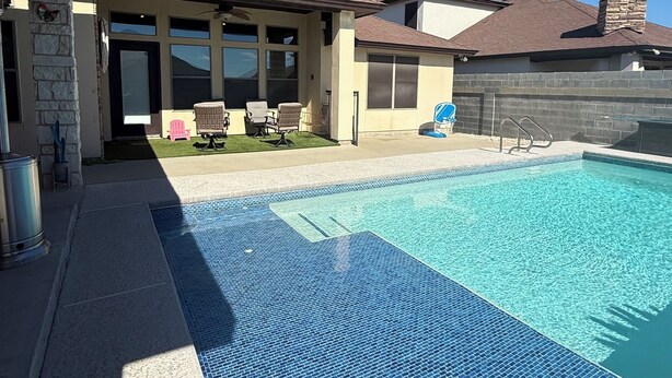 A swimming pool with a tiled deck, a covered patio area with chairs, and a stone pillar.