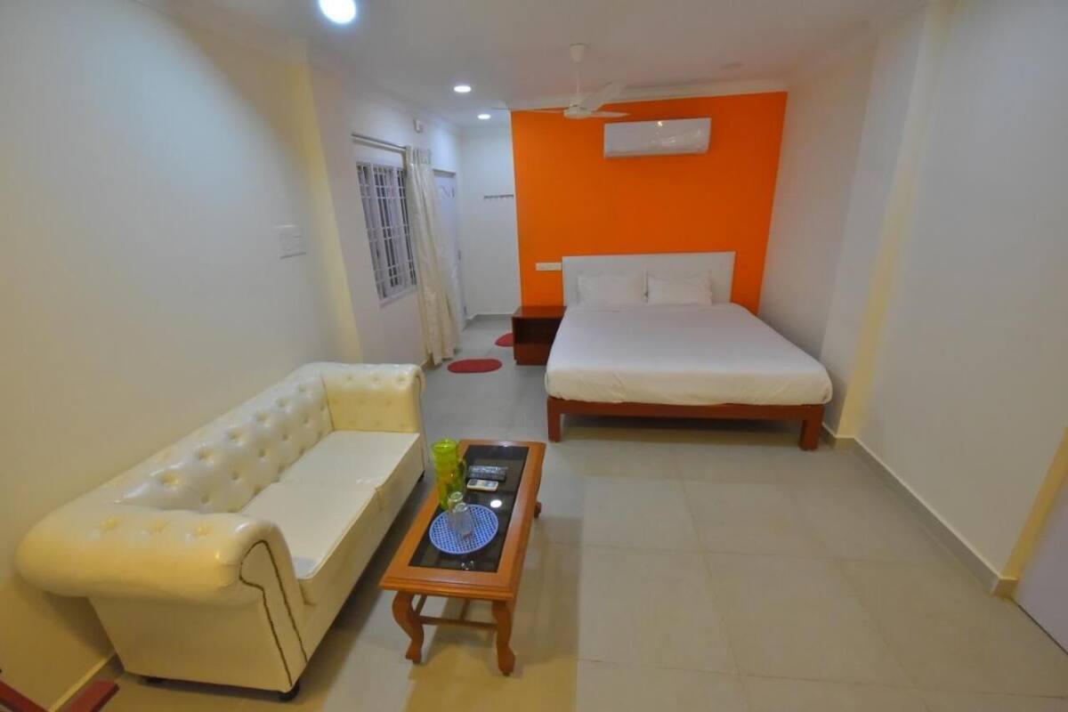 A small, well-lit room with a white sofa, a wooden table with a blue tray, a bed with white bedding, and an orange accent wall.