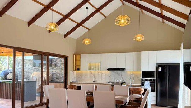 Amplia sala de estar con techos altos, vigas de madera y una cocina moderna con armarios blancos y un refrigerador negro.