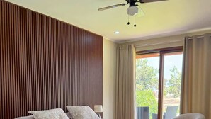 Un dormitorio moderno con una cama grande, paneles de madera en la pared, un ventilador de techo y vistas al exterior a través de un amplio ventanal.