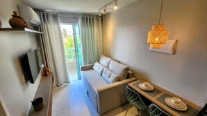 A modern living room with a sofa, a dining table with chairs, a hanging light fixture, and a television on the wall. - Cupe Living - VSH (Ipojuca)