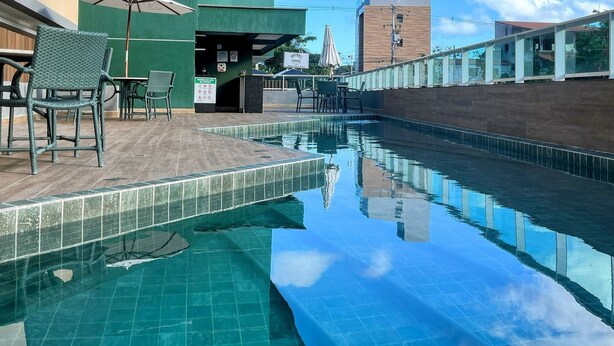 A swimming pool with a tiled edge, outdoor seating with chairs and umbrellas, and a building with a green facade. - Cupe Living - VSH (Ipojuca)