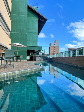 A swimming pool with a tiled edge, outdoor seating with chairs and umbrellas, and a building with a green facade. - Cupe Living - VSH (Ipojuca)