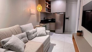 A modern living room with a sofa, a television, and a kitchen area in the background. - Cupe Living - VSH (Ipojuca)