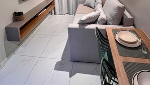 A modern living room with a grey sofa, a flat-screen TV, a wooden shelf, and a dining area with a table set for two. - Cupe Living - VSH (Ipojuca)