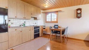 A kitchen with wooden cabinets, a refrigerator, and a dining area with a table and chairs. - Alpine Garden Retreat | Panoramic Mountain Views (Reichenbach im Kandertal)