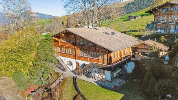 A large wooden chalet with a balcony, surrounded by lush greenery and mountains in the background. - Alpine Garden Retreat | Panoramic Mountain Views (Reichenbach im Kandertal)