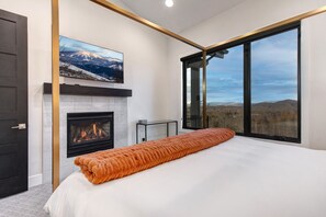 A modern bedroom with a fireplace, a large bed, and a view of the mountains through a large window. - Frost Haven 4 Bedroom Townhouse (Park City)