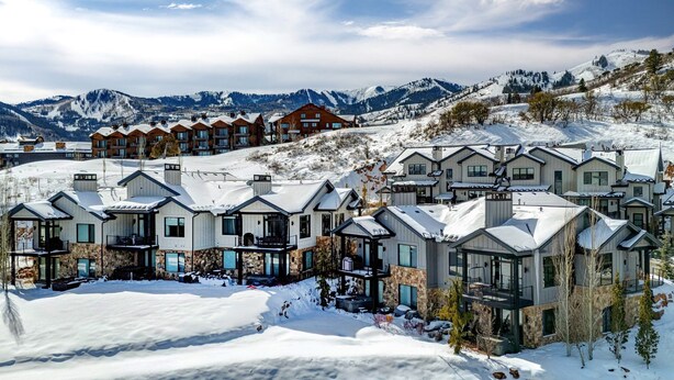 Un área residencial cubierta de nieve con casas modernas, un complejo de esquí al fondo y montañas.