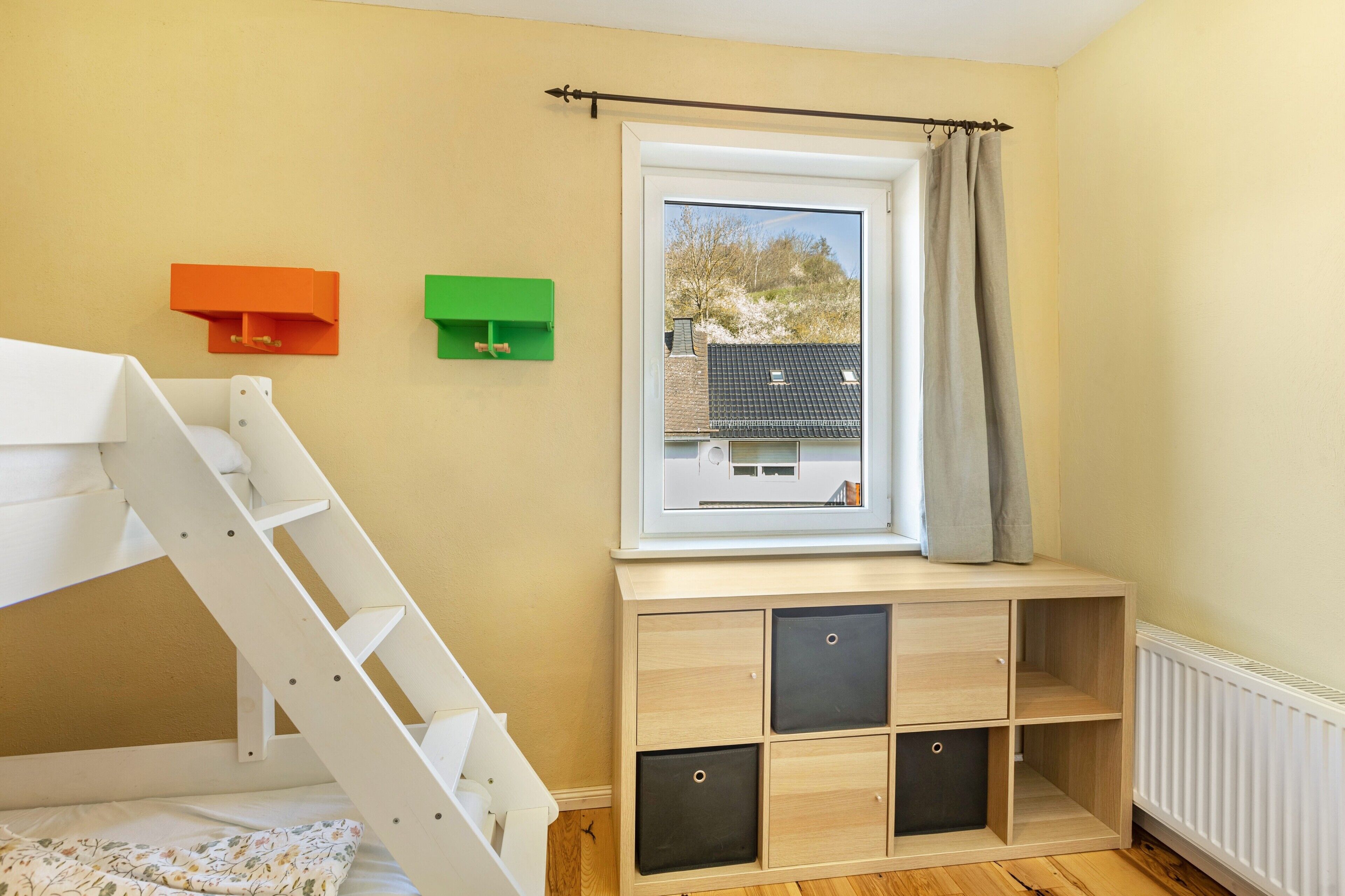 A room with a bunk bed, a window with a view of a house, and a wooden cabinet with drawers.
