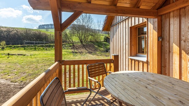 Eine Holzterrasse mit Tisch und Stühlen, mit Blick auf eine Wiese mit Bäumen und eine ferne Hügellandschaft.