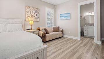 A bedroom with a large bed, a nightstand, a lamp, a beige sofa, and a painting.
