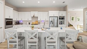 A modern kitchen with white cabinetry, a central island with bar stools, stainless steel appliances, and a dining area with a beige sofa. - Beach House - Magical Purple Haze (Mexico Beach)