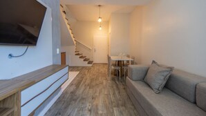 A modern living room with a wooden floor, a grey sofa, a TV mounted on the wall, and a staircase leading to another room.