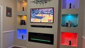Una sala de estar con un televisor montado mostrando un partido de baloncesto, estantes con adornos y una chimenea.