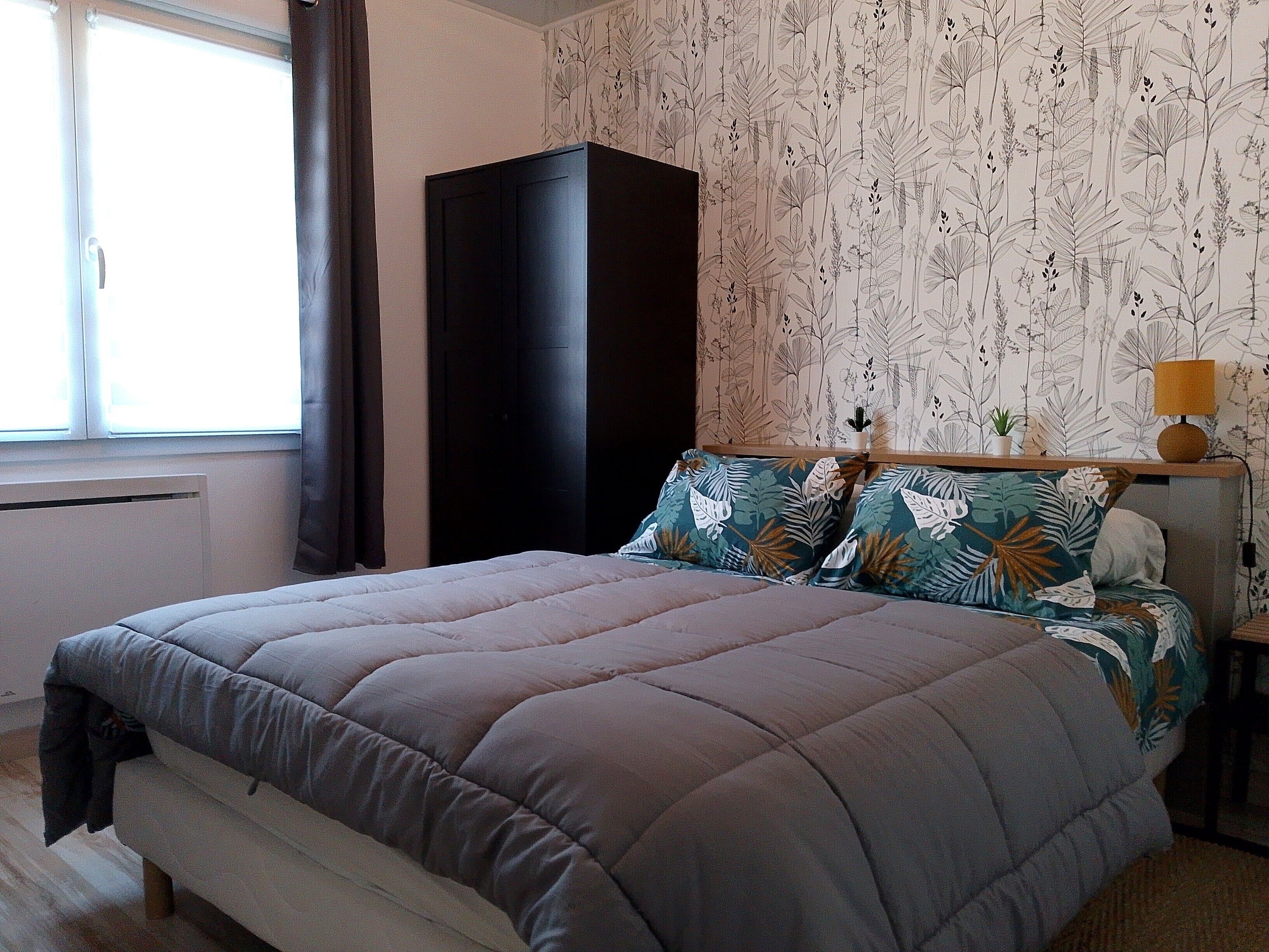 Ein Schlafzimmer mit Bett, Kleiderschrank, Fenster und einer Wand mit Blätermotiv.