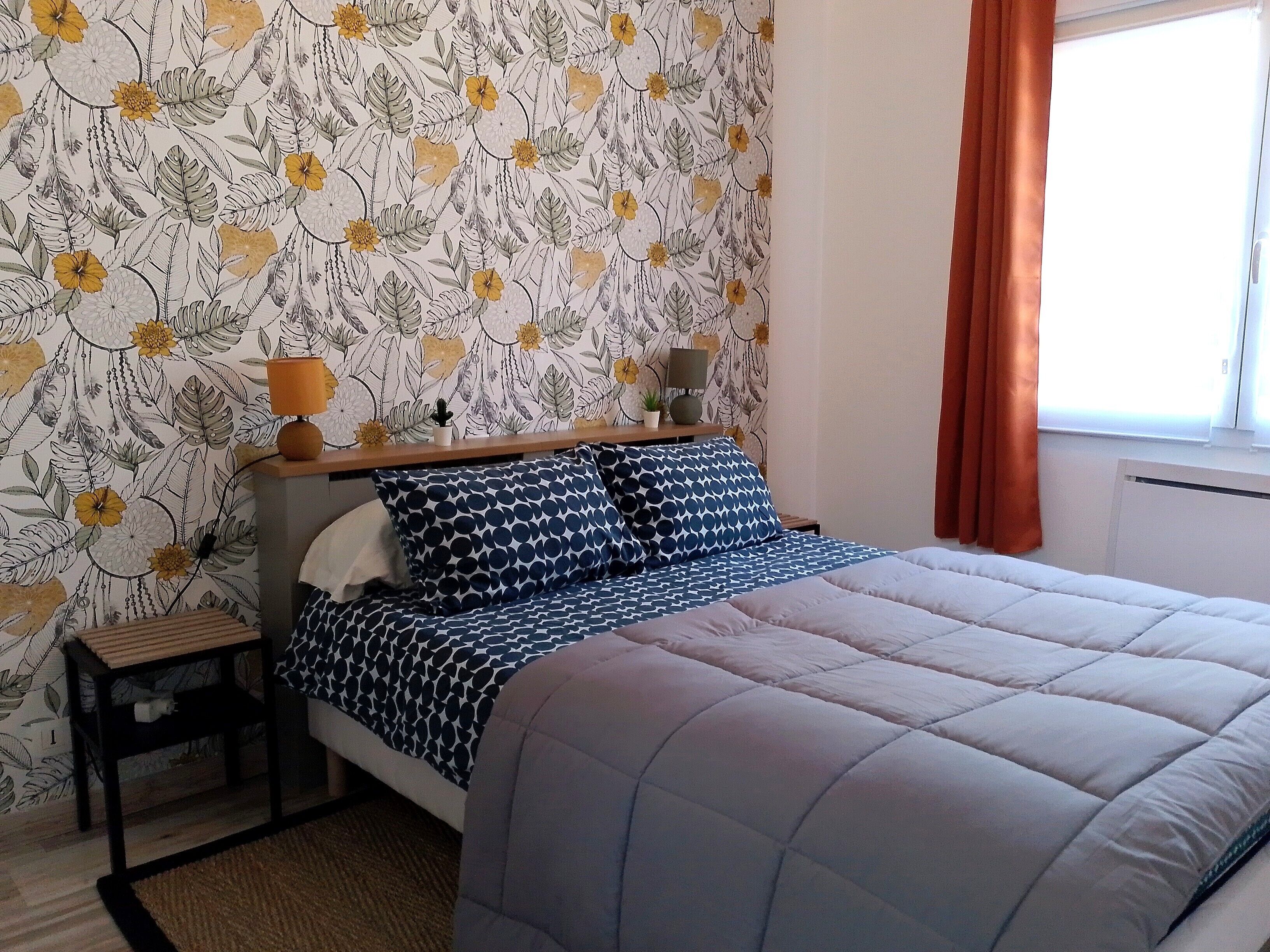 Quarto com papel de parede floral, cama com colcha estampada, mesa de cabeceira com abajur e janela com cortinas.