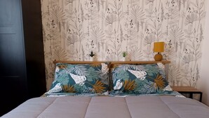Een bed met een witte quilt, twee kussens met een tropisch bladmotief, een houten hoofdbord, een klein nachtkastje met een lamp en een wand met botanische behang.