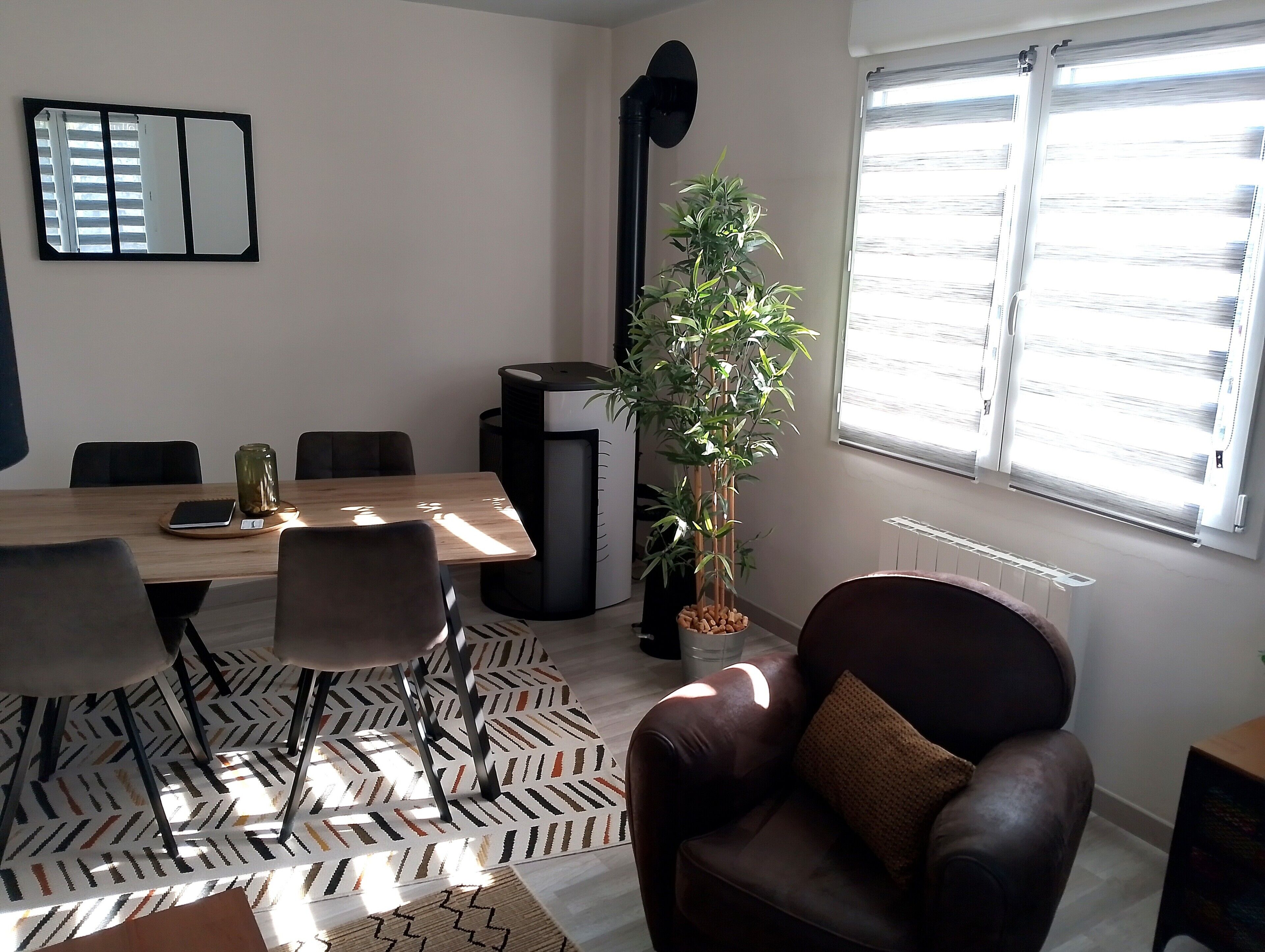 Een moderne woonkamer met een houten eettafel, een bank, een potplant en een groot raam met jaloezieën.