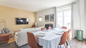 A modern living room with a dining area, a flat-screen TV, and a large window with curtains. - Le Sarrail - Hypercentre & sleeps 12 (Reims)