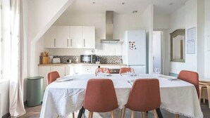 A dining area with a table set for four, white chairs, and a kitchen in the background. - Le Sarrail - Hypercentre & sleeps 12 (Reims)