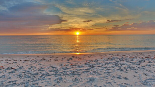 Una playa con huellas que conducen al agua, un atardecer y un cielo despejado.