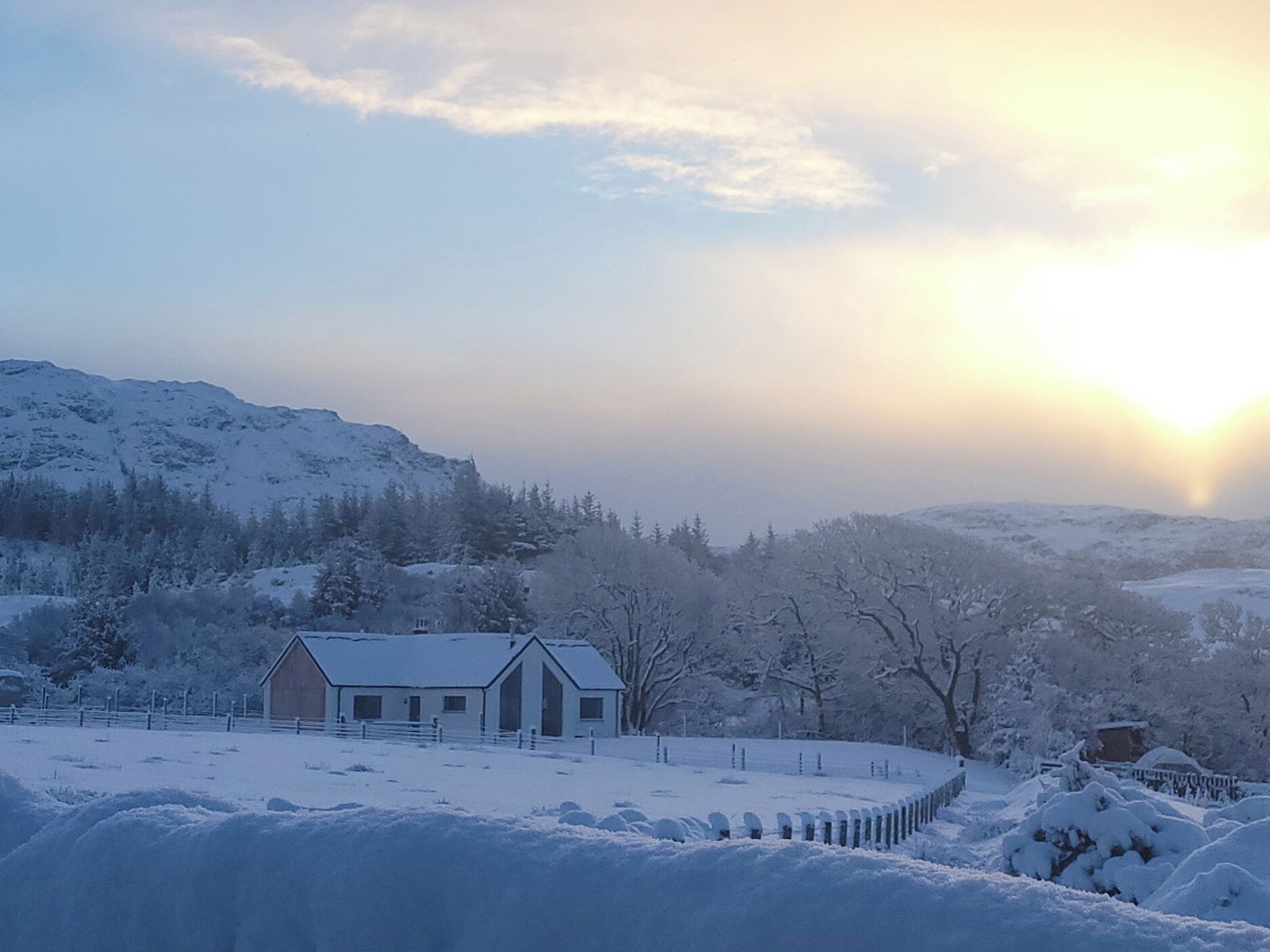 A snow-covered landscape with a house, a fenced area, and a mountain in the background.