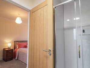 A bedroom with a wooden wardrobe, a bed with red bedding, a nightstand with a lamp, and a glass shower door.