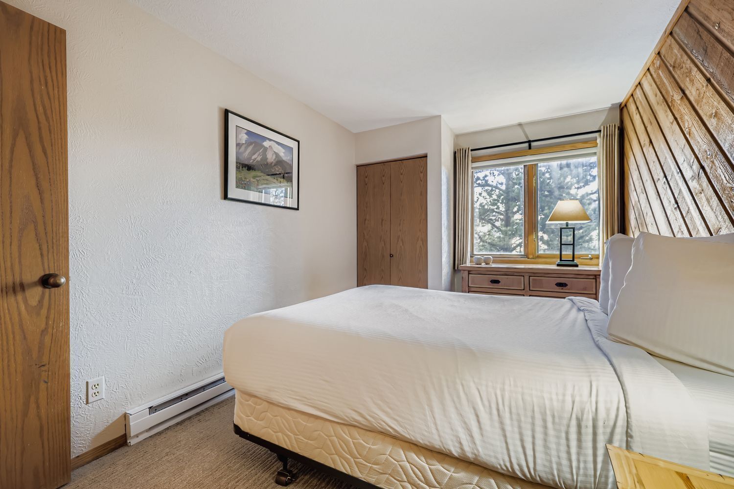 Une chambre à coucher avec un lit, une fenêtre donnant sur un beau paysage, une table de chevet et une porte en bois.