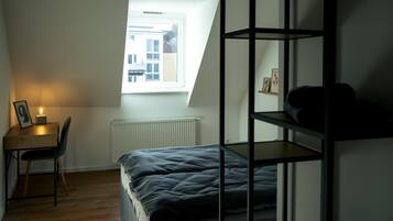 A dormitory room with a bunk bed, a desk with a chair, a window, and a radiator.