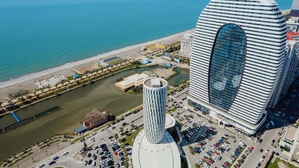 A modern hotel with a distinctive curved facade and a central cylindrical tower, located near a waterfront with a parking area and a bridge. - Studio by Alliance (Batumi)