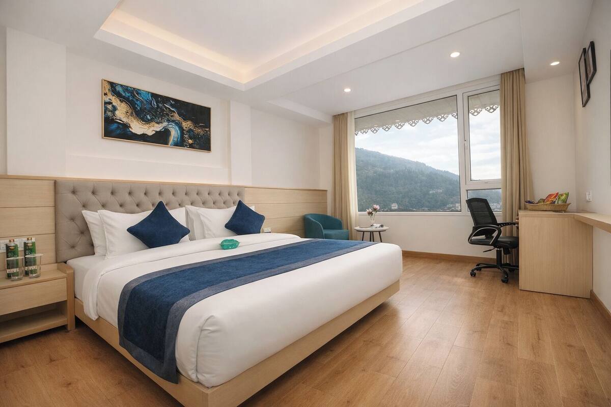 A hotel room with a large bed, a desk, a chair, a window with a mountain view, and a painting on the wall.