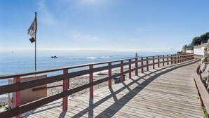 Ein Holzsteg führt zu einem Strand mit einer Fahnenmast und wolkenlosem, blauem Himmel.