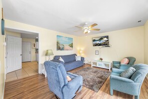 Ein Wohnzimmer mit einem blauen Sofa, zwei Sesseln, einem Couchtisch, einem Teppich und einem Deckenventilator.