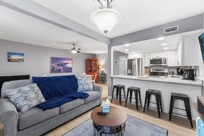 Ein modernes Wohnzimmer mit grauem Sofa, einem Couchtisch und einer Küchenzeile mit Bar und Hockern.