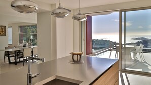Une cuisine moderne dotée d’un grand îlot central, de luminaires suspendus et offrant une vue sur la mer grâce à de grandes fenêtres.