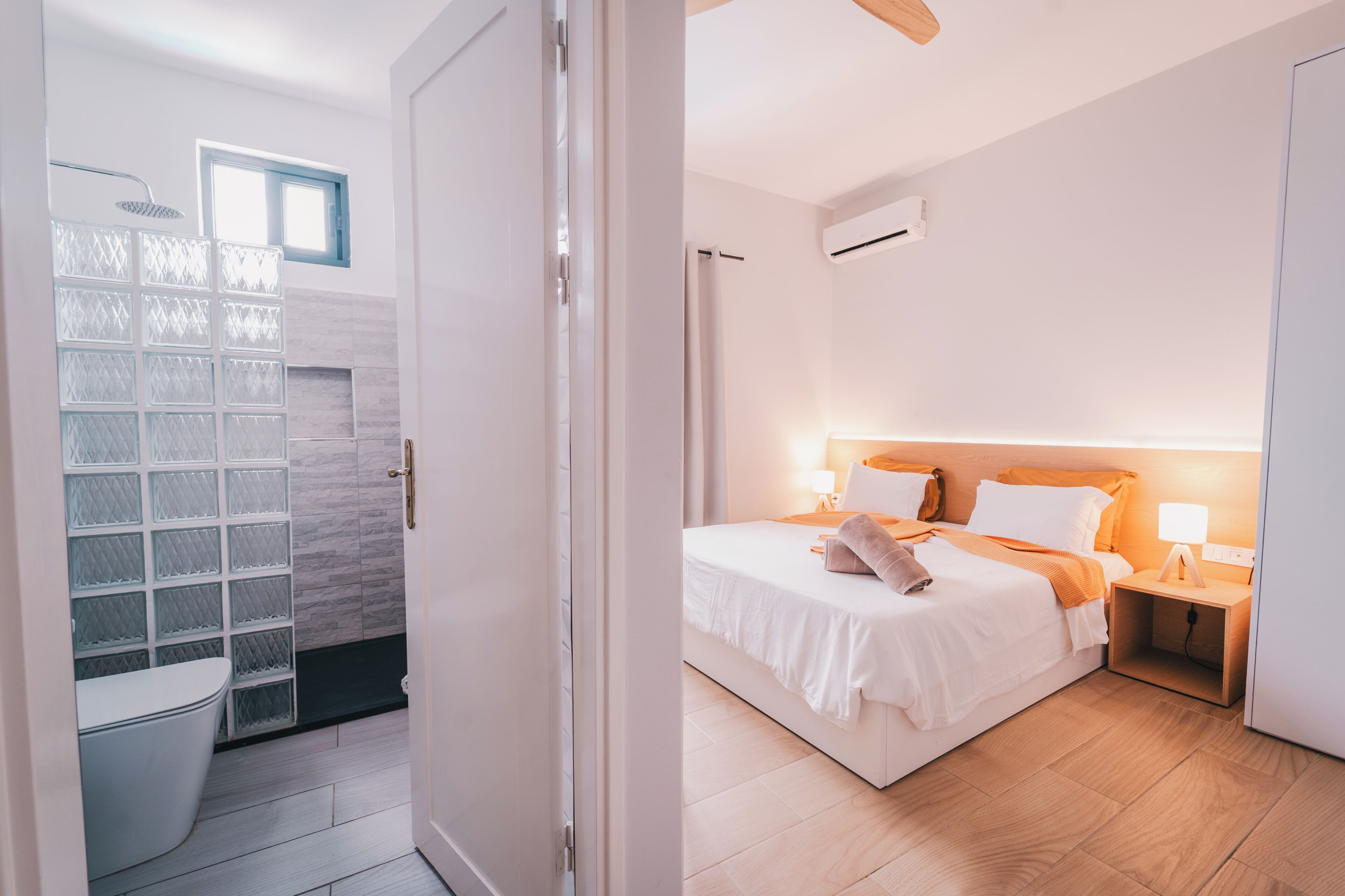 A modern hotel room with a shower, a bed with white bedding, a wooden floor, and a nightstand with a lamp.