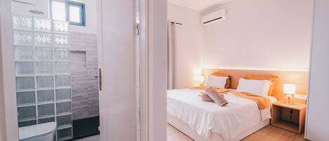 A modern hotel room with a shower, a bed with white bedding, a wooden floor, and a nightstand with a lamp.