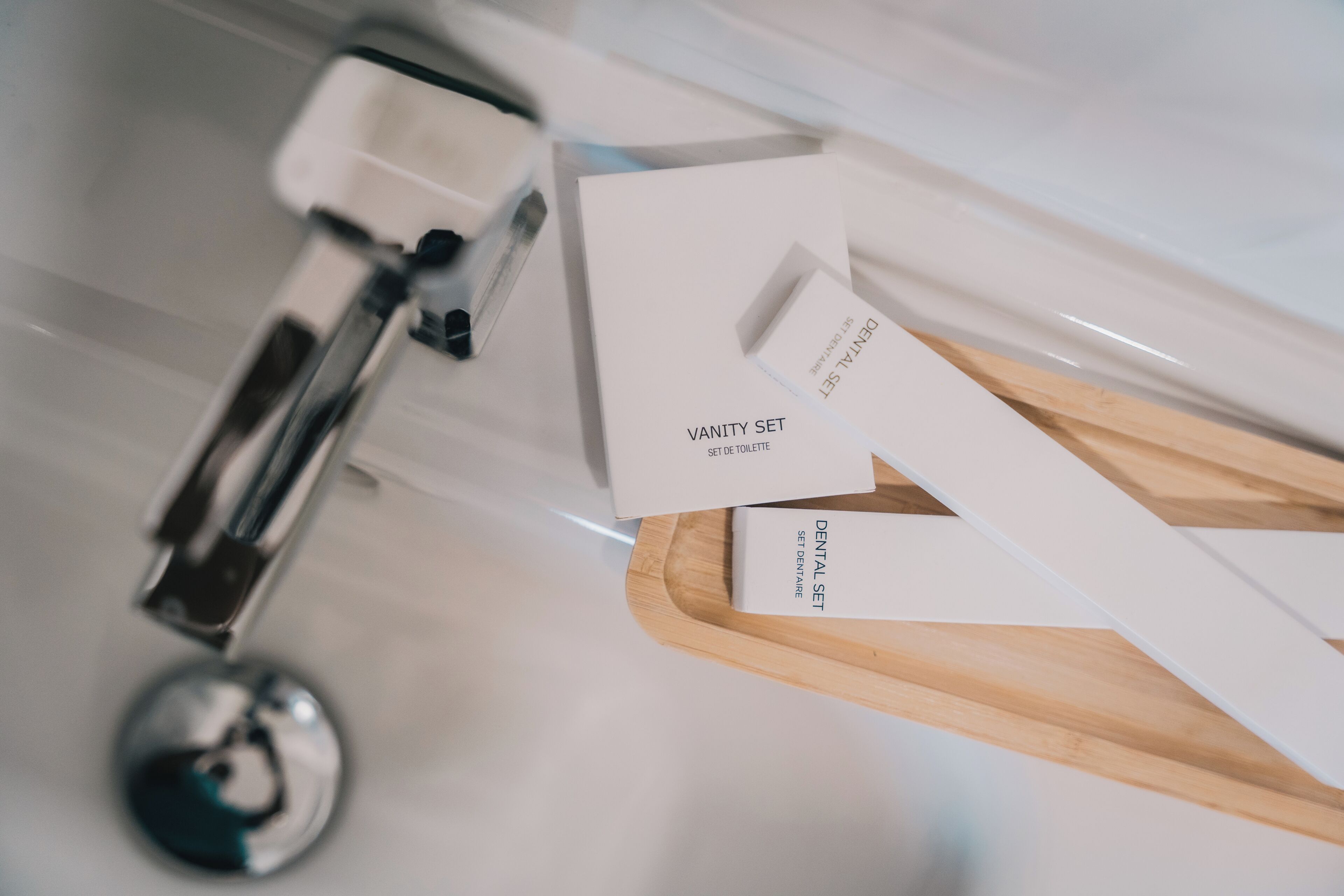 A bathroom with a modern faucet, a wooden tray, and two boxes labeled 'VANITY SET SET DE TOILETTE' and 'DENTAL SET.'