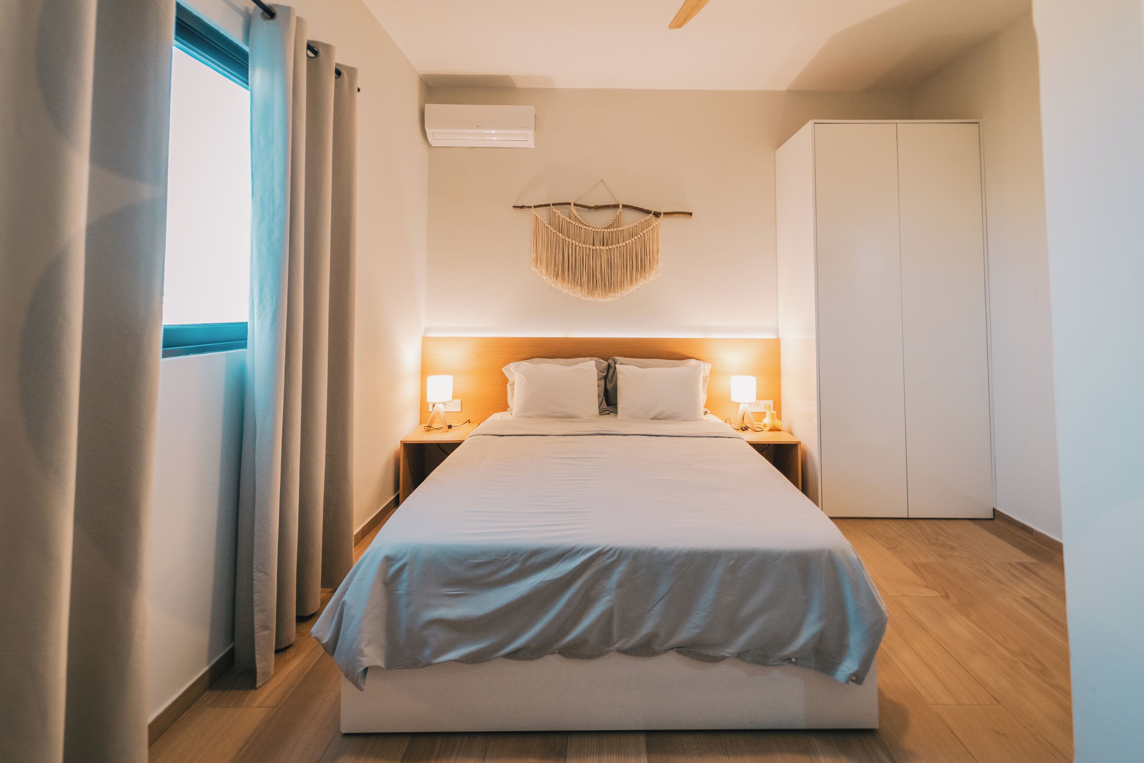 Una habitación de hotel con una cama, lámparas de noche, un armario y un tapiz de macramé en la pared.