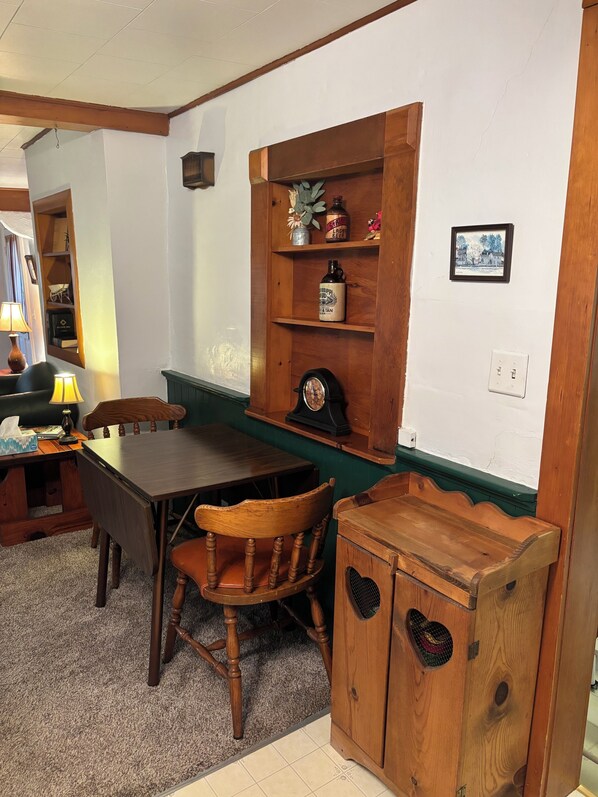 A cozy room with a wooden table, chairs, a small cabinet, and a shelf with various items.
