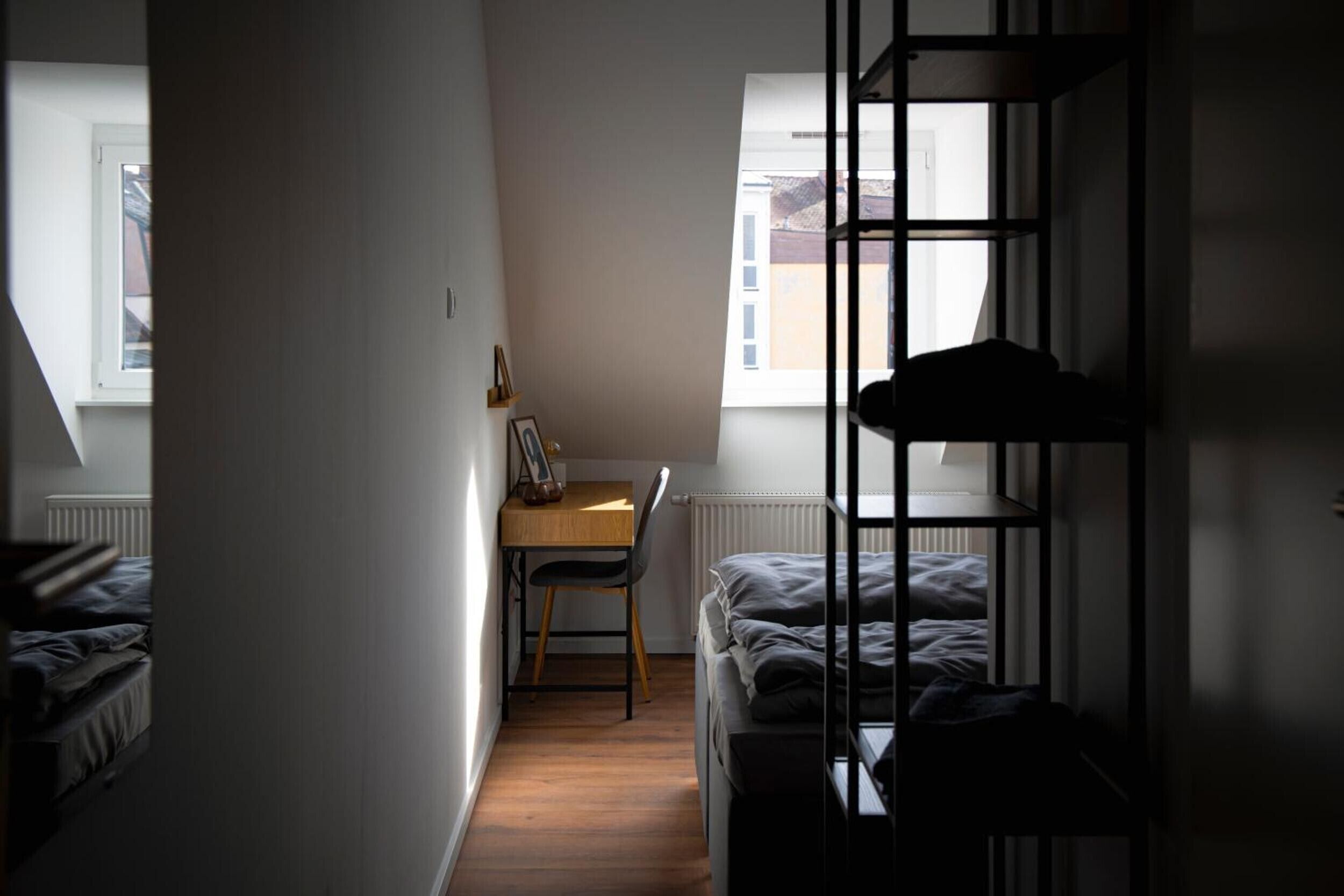 A narrow room with a bed, a desk, and a chair. There is a window providing natural light.