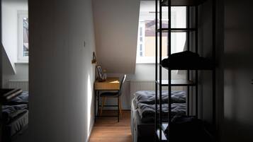 A narrow room with a bed, a desk, and a chair. There is a window providing natural light.