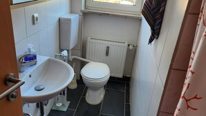 A small bathroom with a white sink, toilet, and radiator. A window provides a view of buildings outside. - Einfache 1-Zimmer Wohnung (Bad Woerishofen)