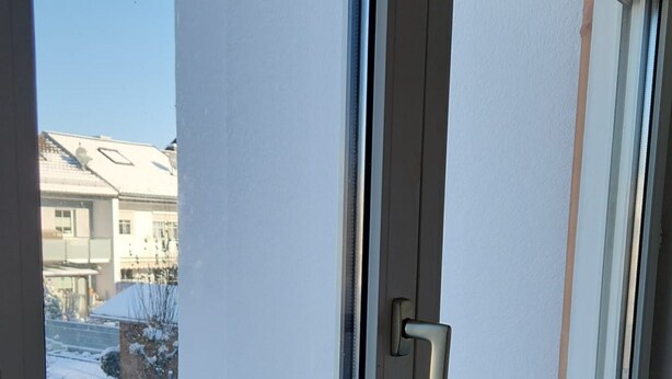 A window with a white frame and a door handle, overlooking a snowy street with buildings. - Einfache 1-Zimmer Wohnung (Bad Woerishofen)