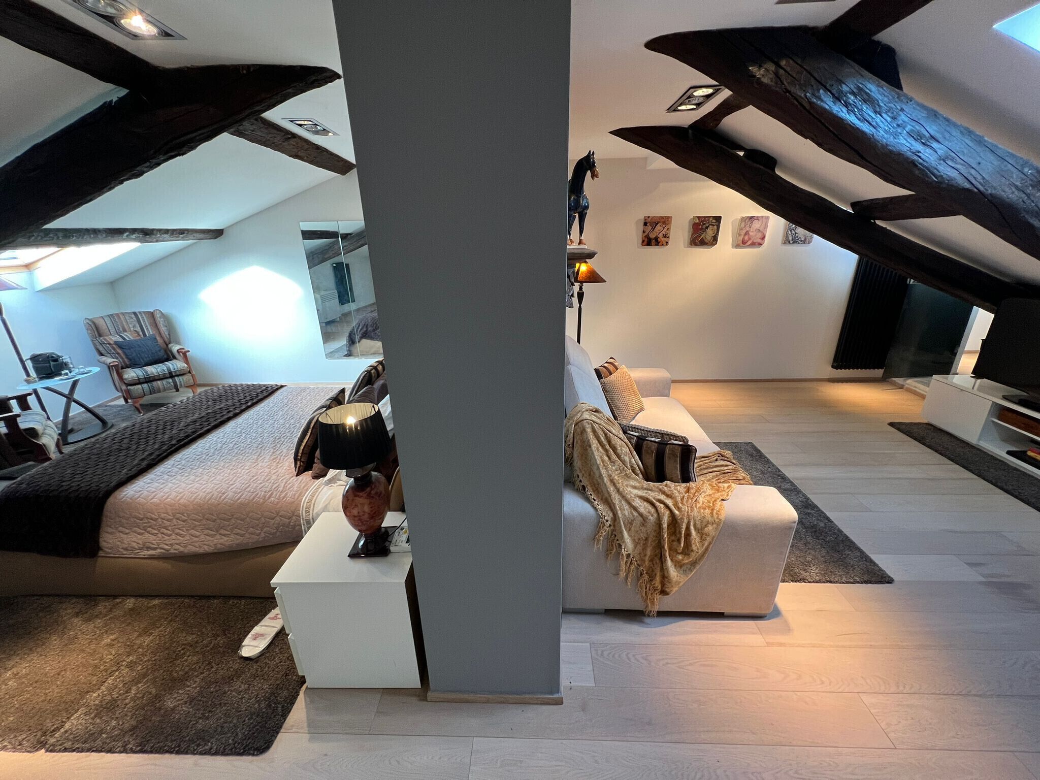 A modern loft bedroom with a high ceiling, exposed beams, a bed, a sofa, and a small table.