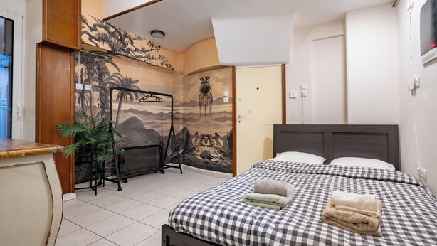 A hotel room with a checkered bedspread, a wooden headboard, a wall mural, and a desk with a plant. - Kolonaki Cozy (Athina)